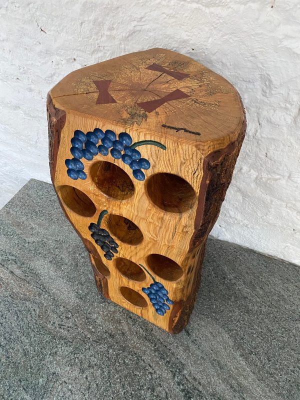 Top over view of this English Ash Tree Stump wine rack showing 3 bow ties in Padauk wood on the top end grain helping to stabilise the splits, bark still attached to the sides and back and 3 bunches of grapes engraved onto the front painted in blue and black, handcrafted by Crystal Woods