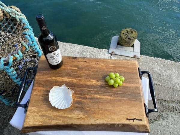 Overview shot of this sycamore chopping board with black metal handles displayed on a waterfront with a bottle of wine and grapes