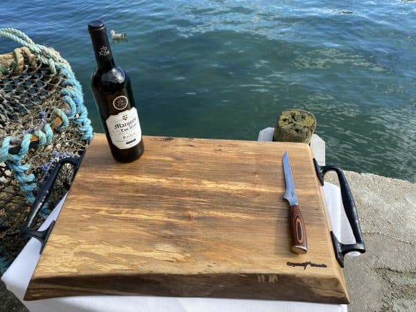 Side overview of this sycamore chopping board with black metal handles displayed on a waterfront with a bottle of wine and knife