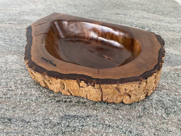 Angled view of this Handmade English Walnut Sculptured Reservoir Bowl with live edge detail, created by Crystal Woods, finished in danish oil, branded with their logo stamp