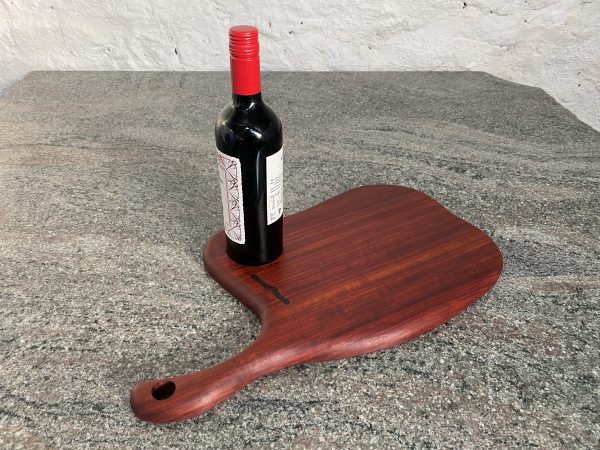 Slender Curved Meat Cleaver Style Charcuterie Board in Exotic Padauk Wood, with a scolloped indent to take a glass or wine bottle, showing a bottle of wine on the serving board, handcrafted by Crystal Woods