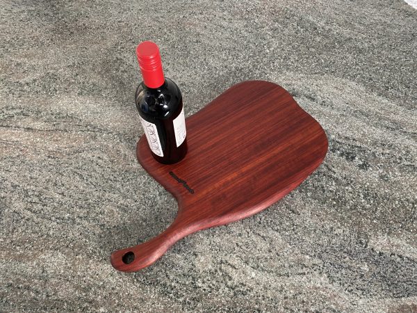 Birds eye view of this Slender Curved Meat Cleaver Style Charcuterie Board in Exotic Padauk Wood, with a scolloped indent to take a glass or wine bottle, showing a bottle of wine on the serving board, handcrafted by Crystal Woods and branded with their logo stamp