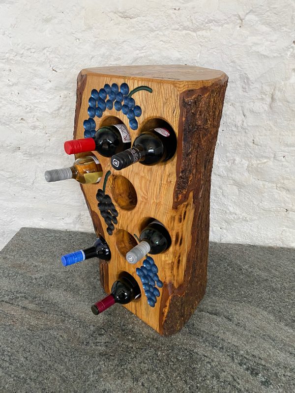 8 bottle small tree stump wine rack made from English Ash locally sourced in Devon, with 3 bunches of grapes engraved on the front painted in blue and black, displaying 6 bottles of wine in the inserted holes, finished in multiple coats of oil, handcrafted by Crystal Woods